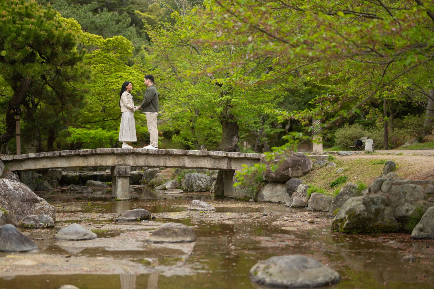 Couple standing on a stone bridge in a lush Japanese garden with green trees and a tranquil stream.