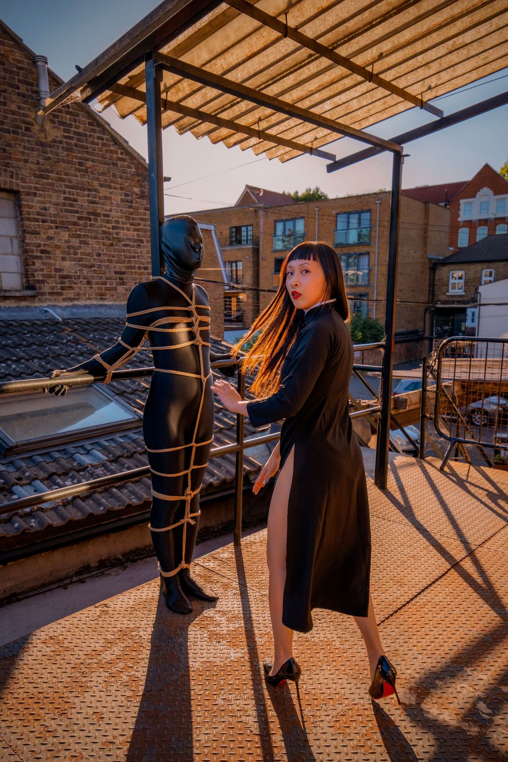 Woman in black dress stands on rooftop terrace next to a rope-bound figure in black bodysuit at dusk.