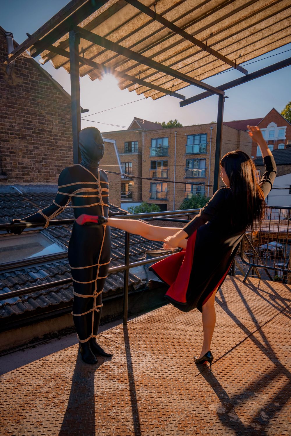 Two performers on a rooftop terrace, one in rope bondage costume, one in black dress, posing at sunset.