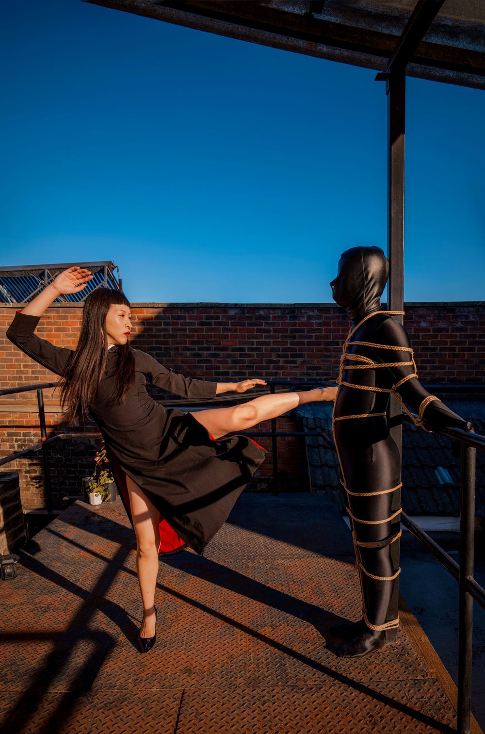 Woman in black dress poses dramatically beside a rope-bound figure on a rooftop terrace against a clear blue sky.