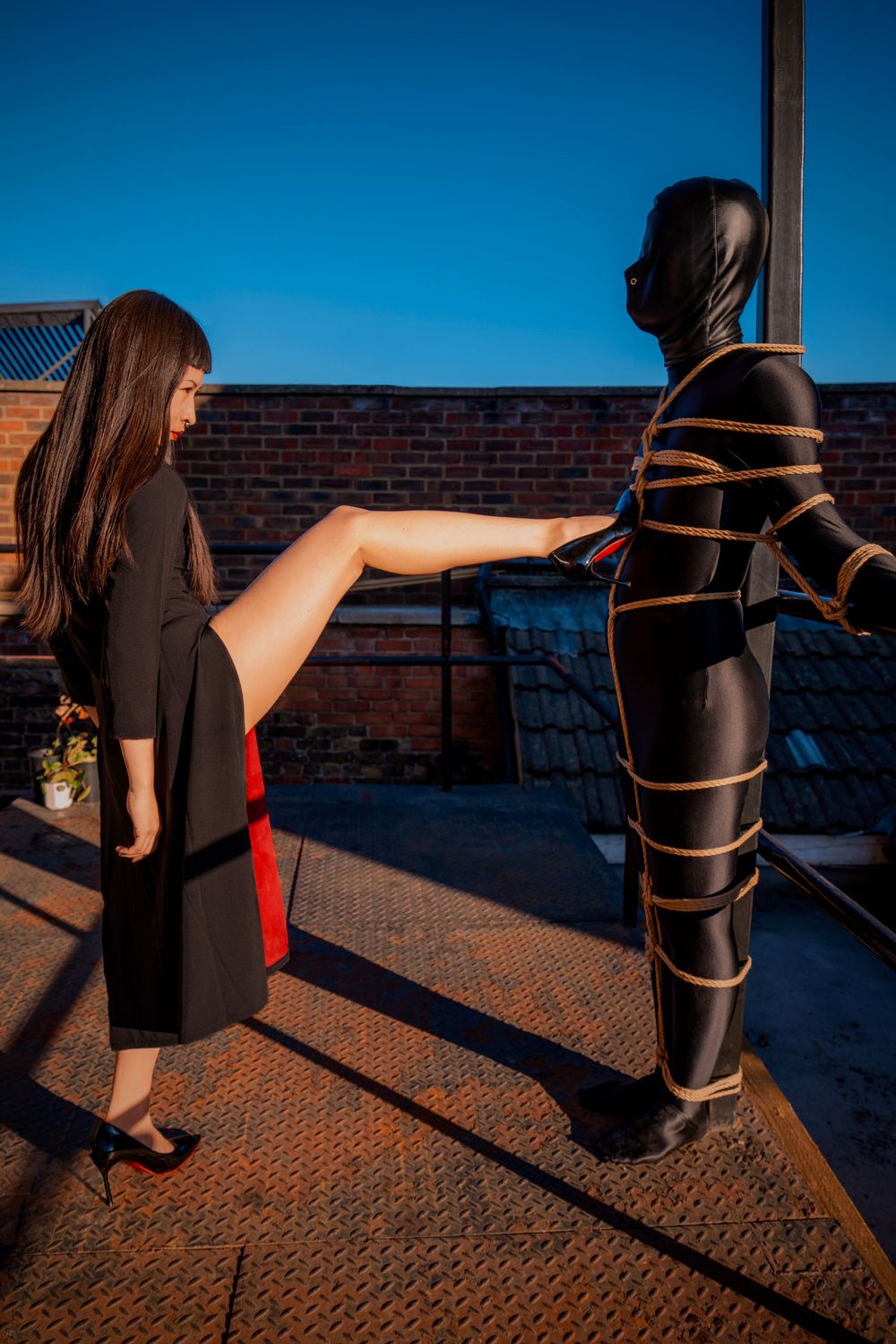 Woman in black dress extends leg toward person in black latex suit tied with rope outdoors on rooftop.
