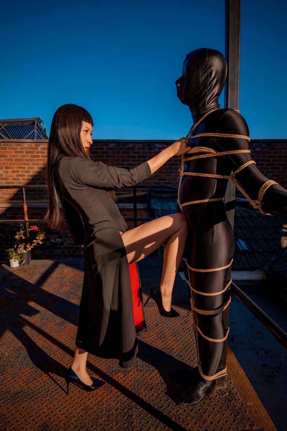 Woman in black coat interacting with person in black latex suit bound with rope on rooftop at dusk.