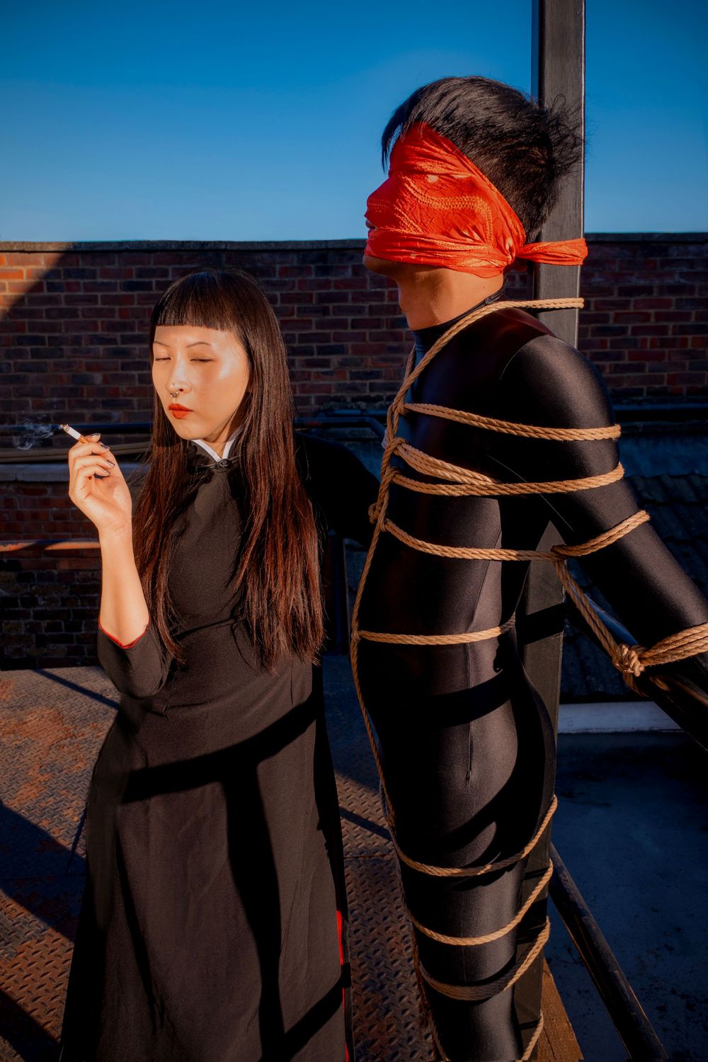 Woman in black dress smoking beside a person bound in rope with red blindfold in shibari art photography.
