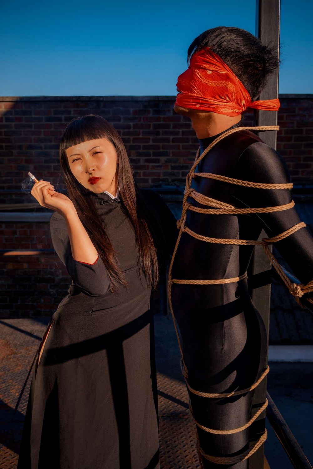 Two people in black outfits on a rooftop, one blindfolded and tied with rope, the other holding a cigarette.
