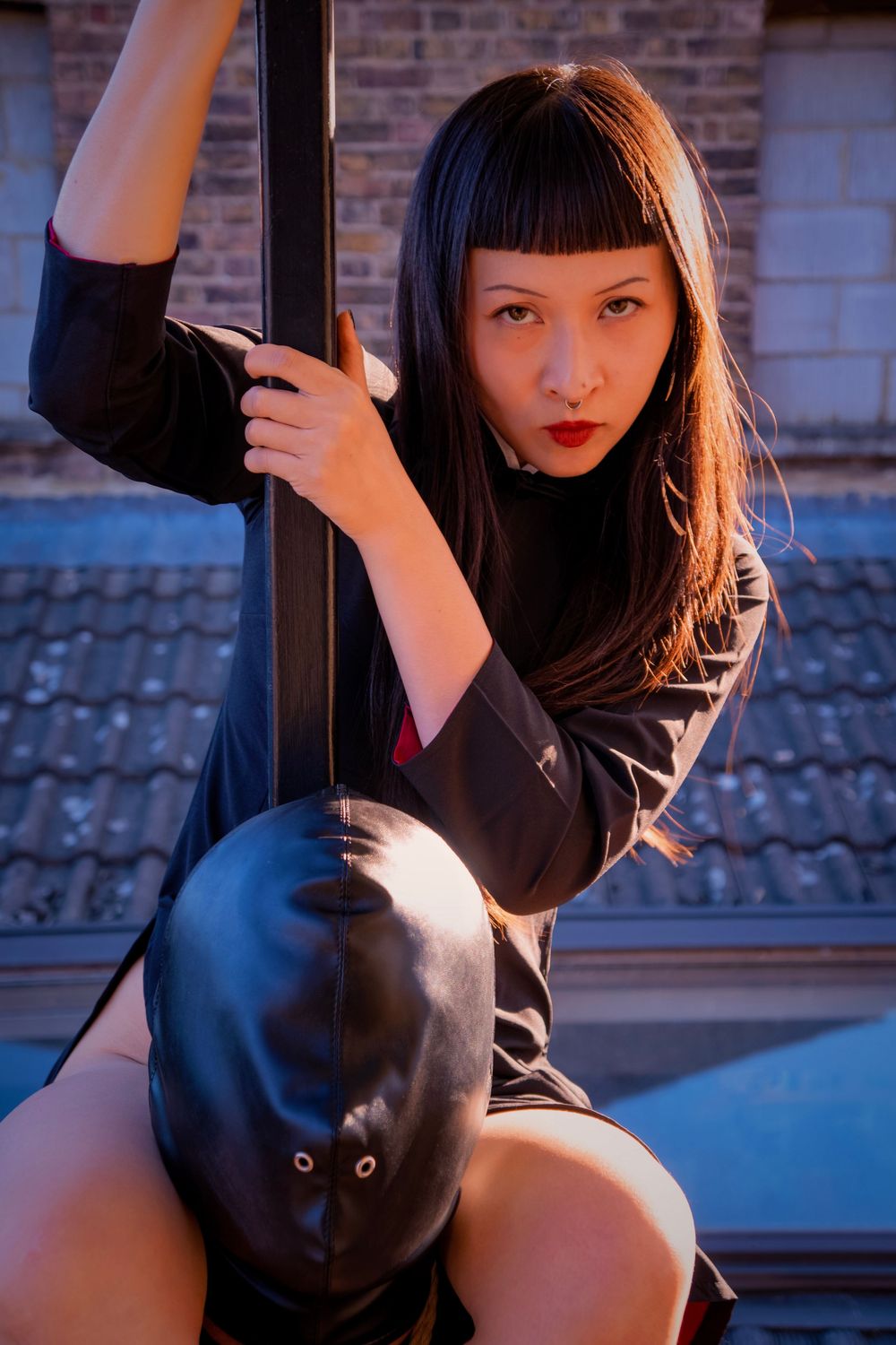 Young woman with dark hair and red lips posing against a metal pole outdoors near a brick building.