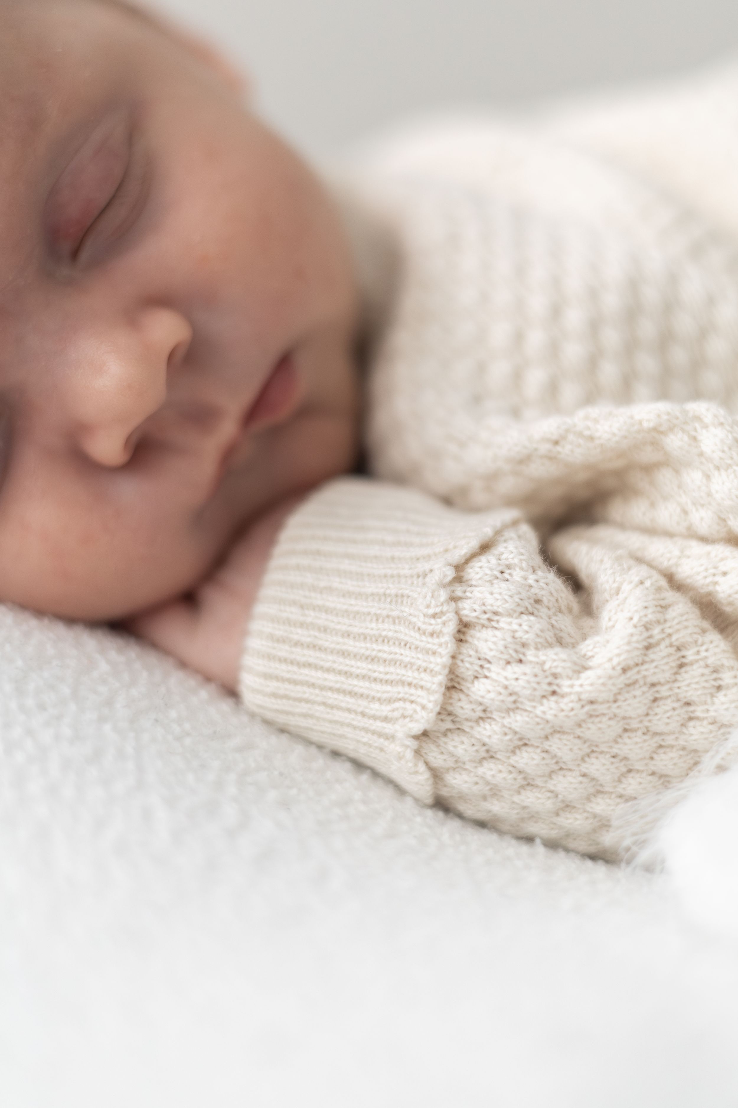 Peaceful newborn baby sleeping in a cream knit onesie on a soft white blanket.