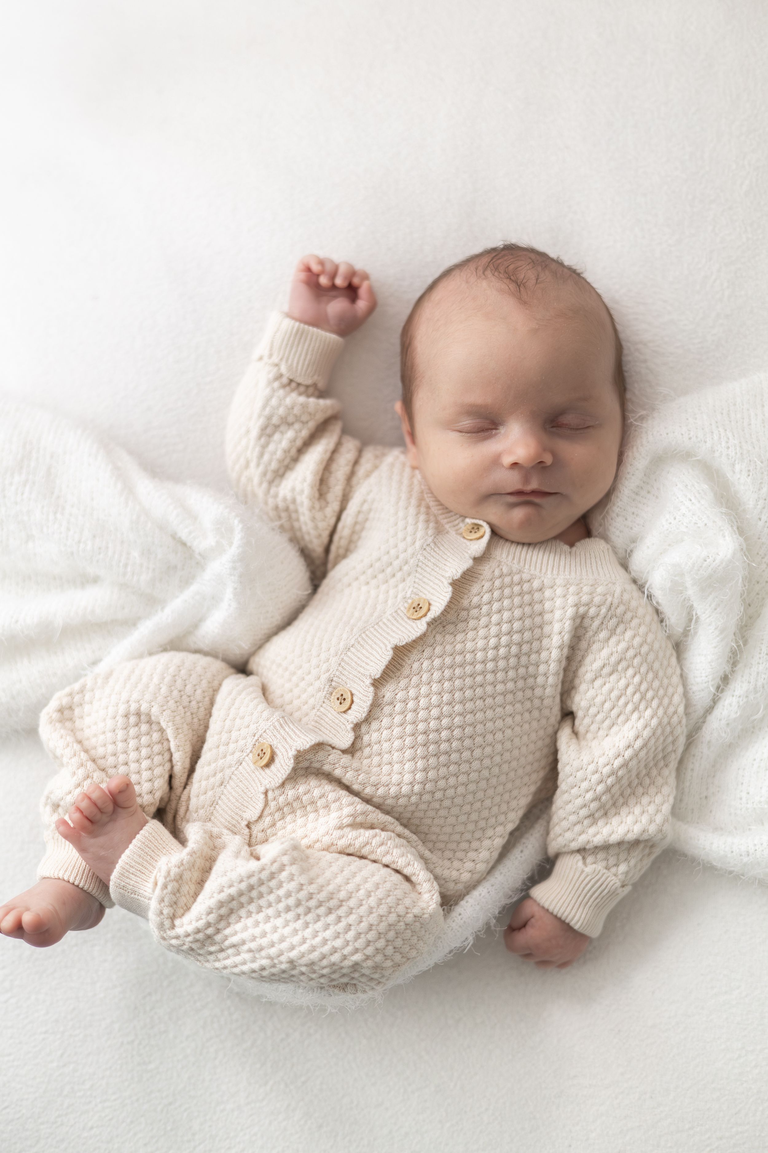 Sleeping newborn baby in a cream knit romper lying on a white blanket with one arm raised.