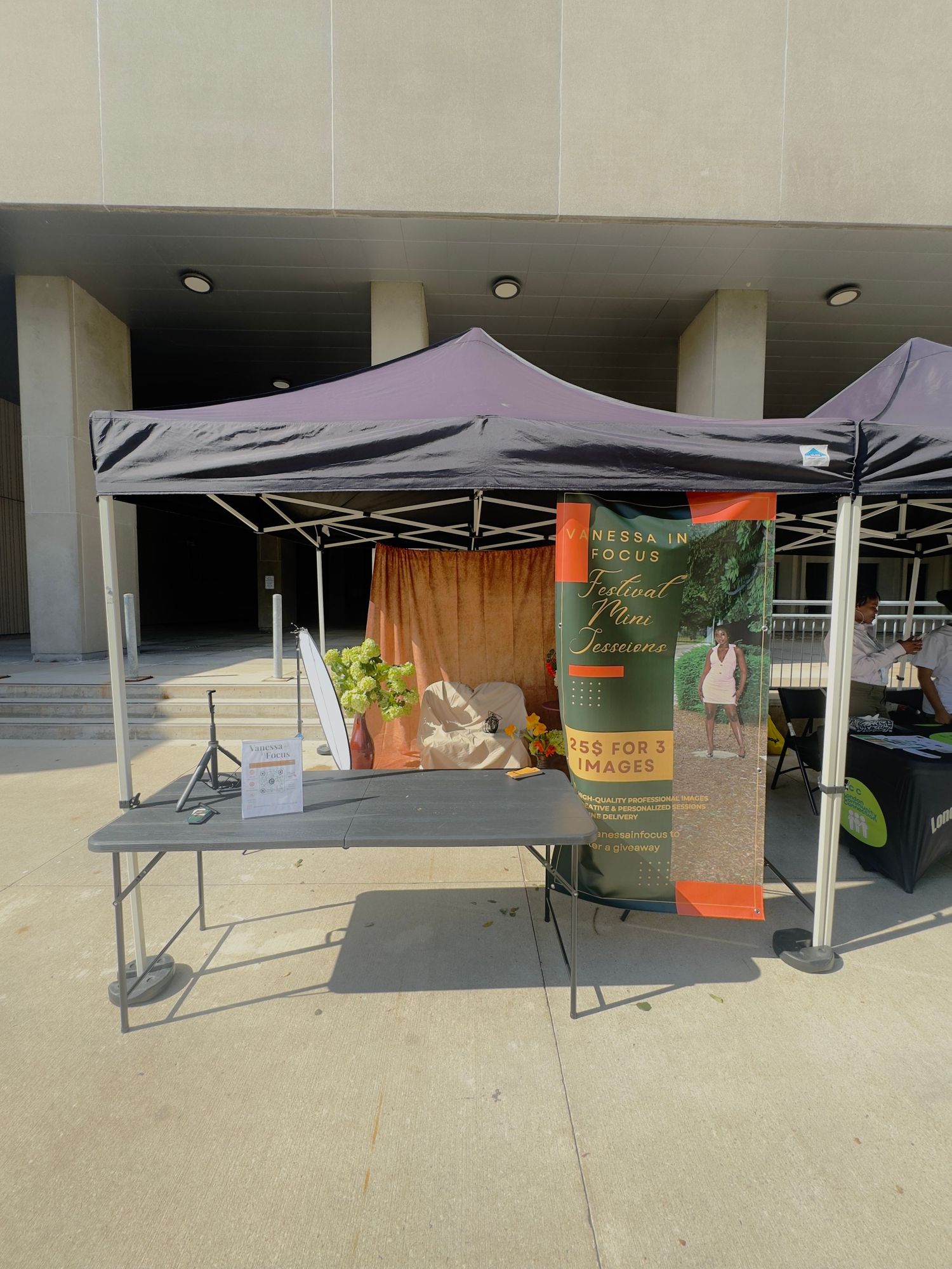 Outdoor vendor booth with black canopy tent, folding table, and promotional banner at an open-air event near a concrete building.