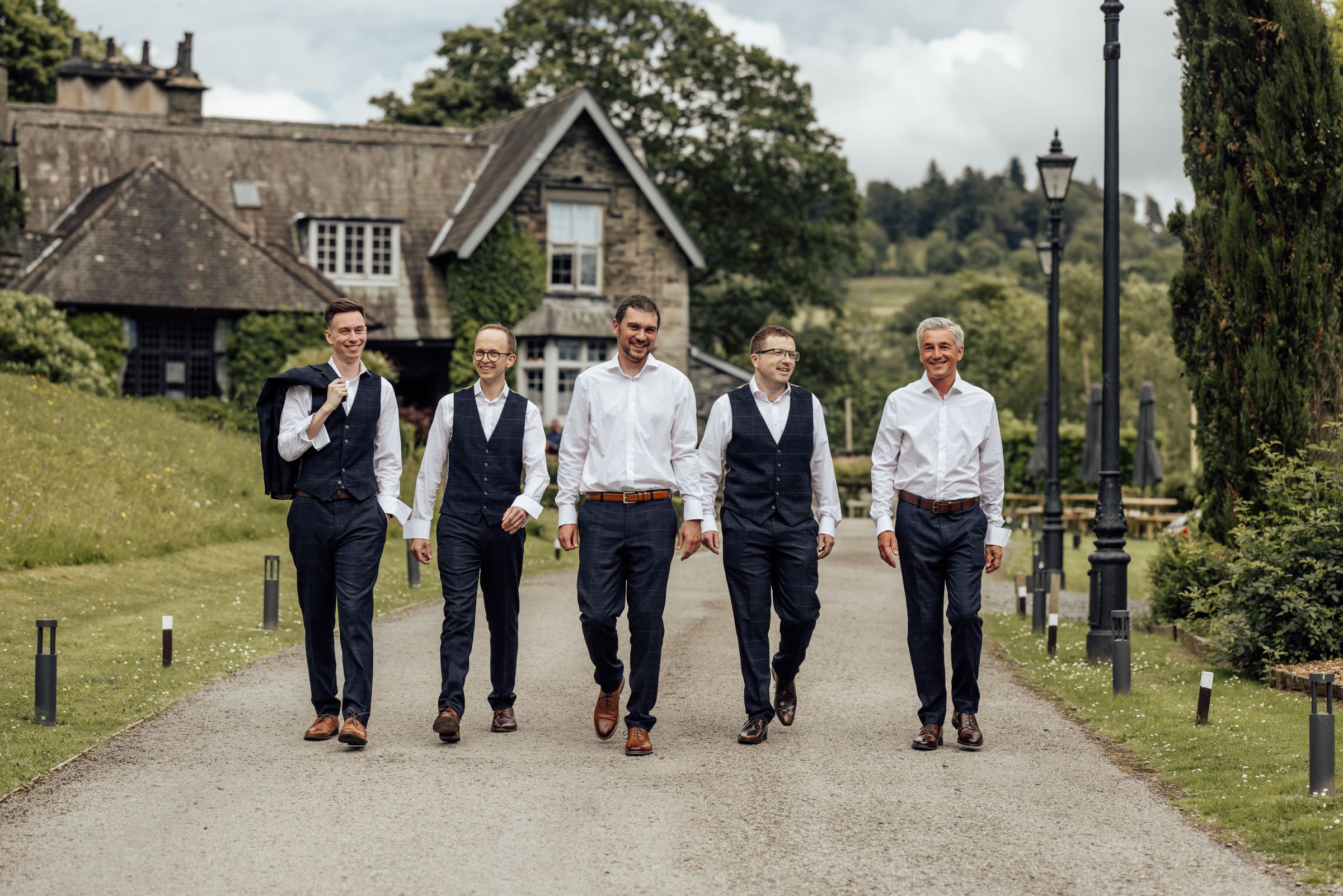 Five groomsmen in navy suits walk confidently down a tree-lined driveway outside a stone country house venue.