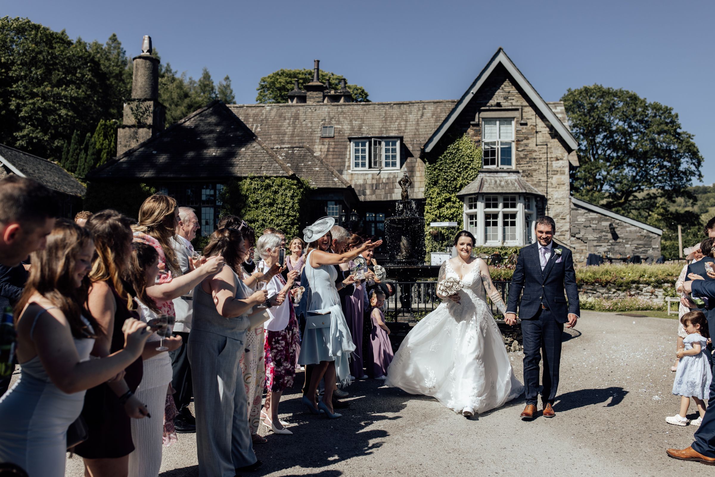 Bride and groom walk through cheering guests throwing confetti outside Broadoaks Country house on a sunny wedding day.