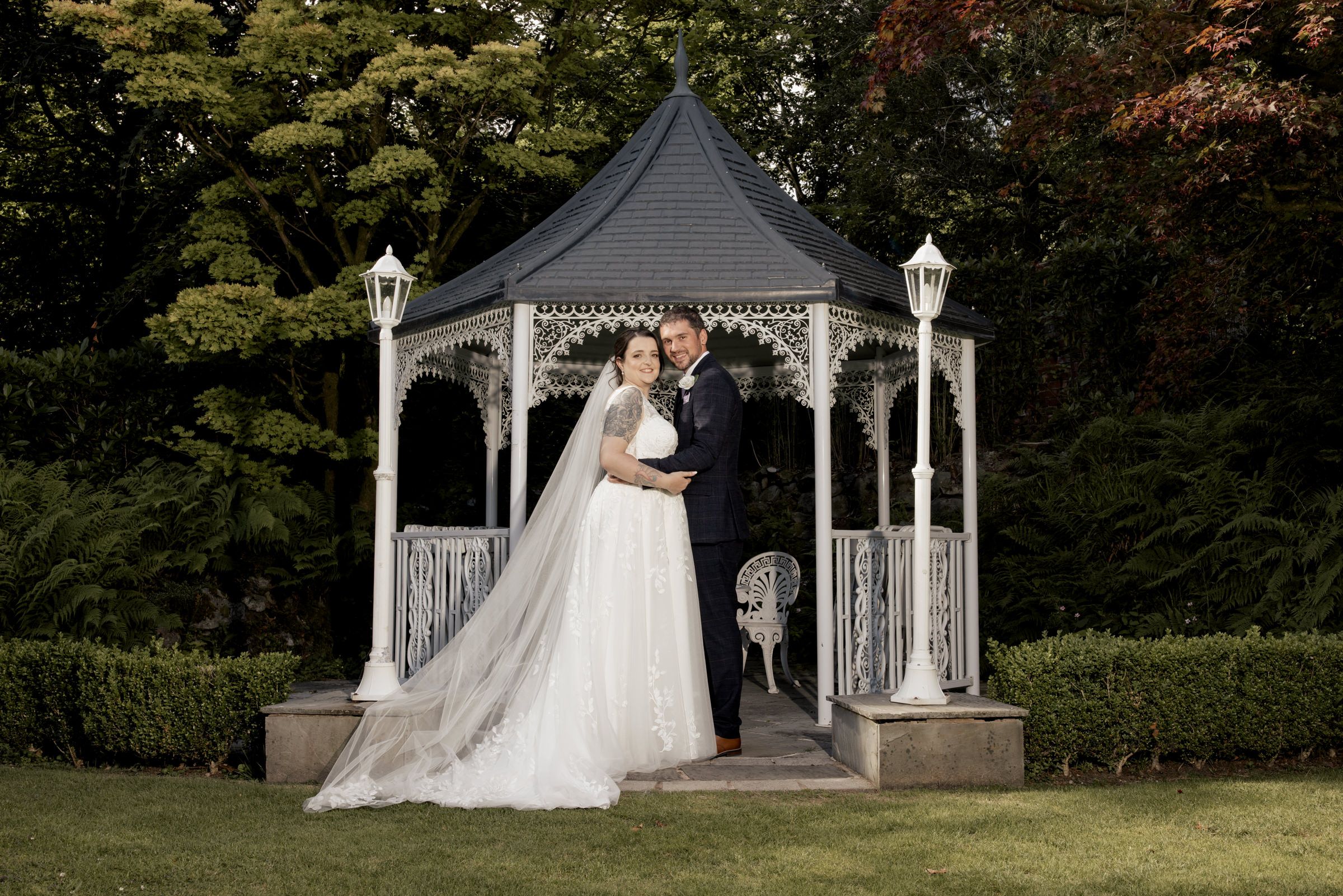 Bride and groom pose romantically under a white gazebo surrounded by lush greenery on their wedding day.