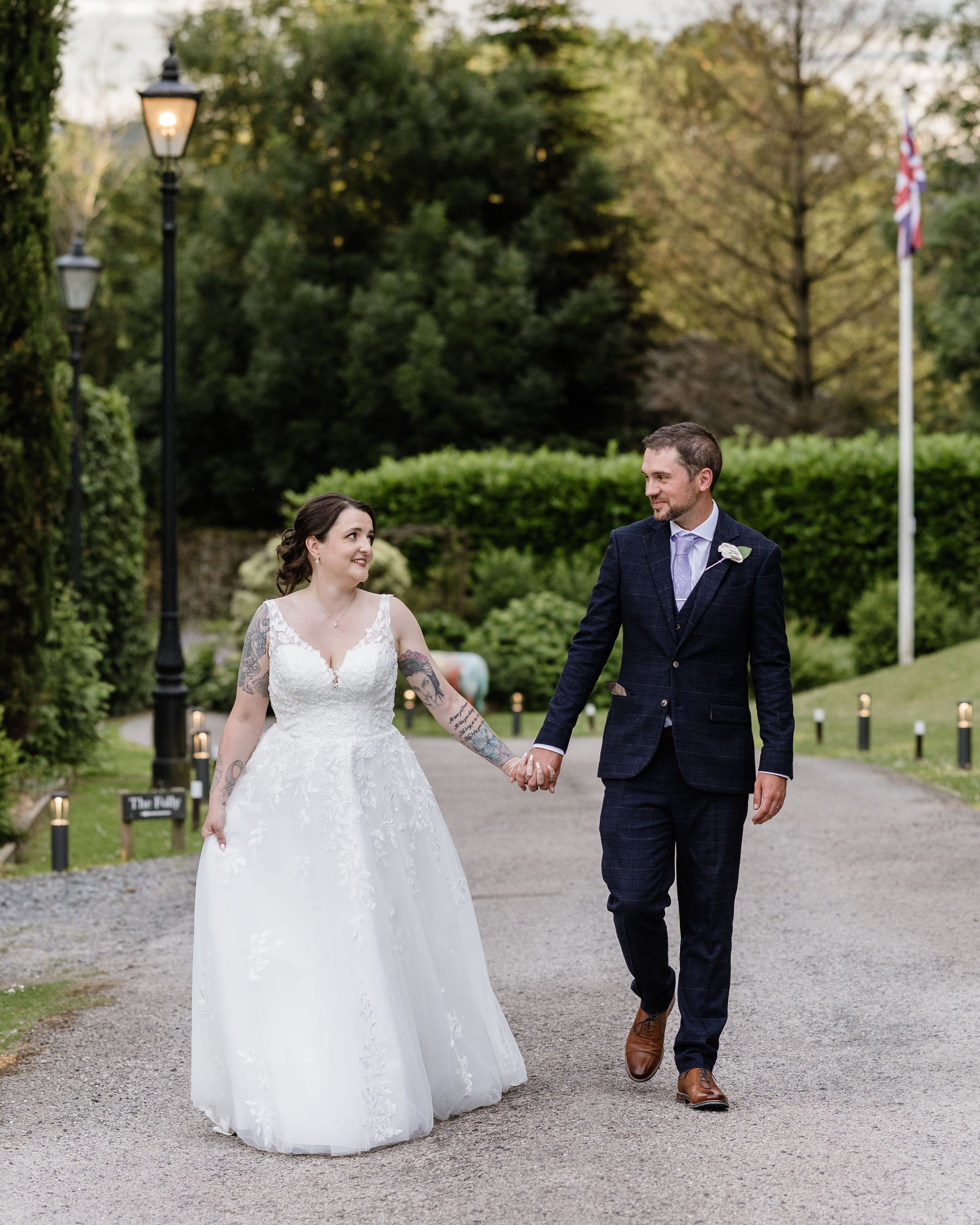 Bride and groom walk hand-in-hand along a tree-lined path, smiling at each other in elegant wedding attire.