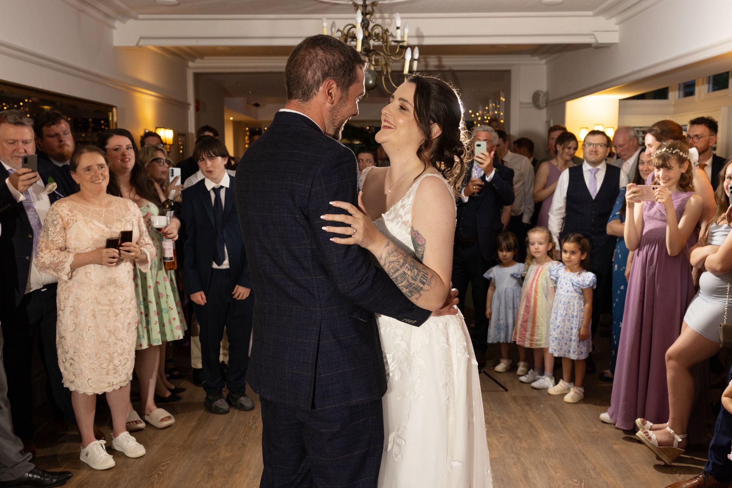 Bride and groom share their first dance surrounded by smiling wedding guests in an elegant reception venue.