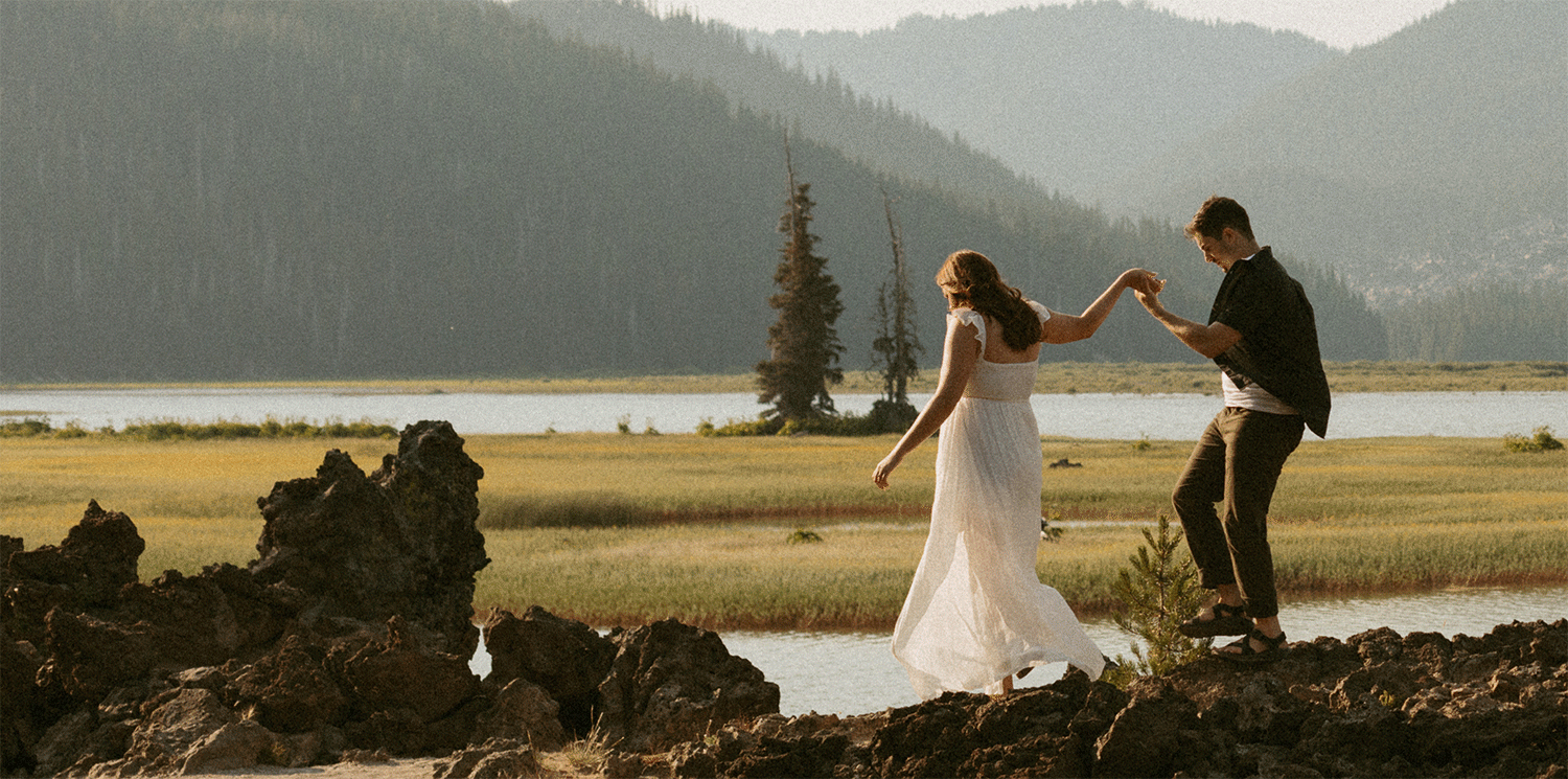 Bride in a flowing white gown holds hands with groom while walking across volcanic rocks beside a mountain lake at sunset.