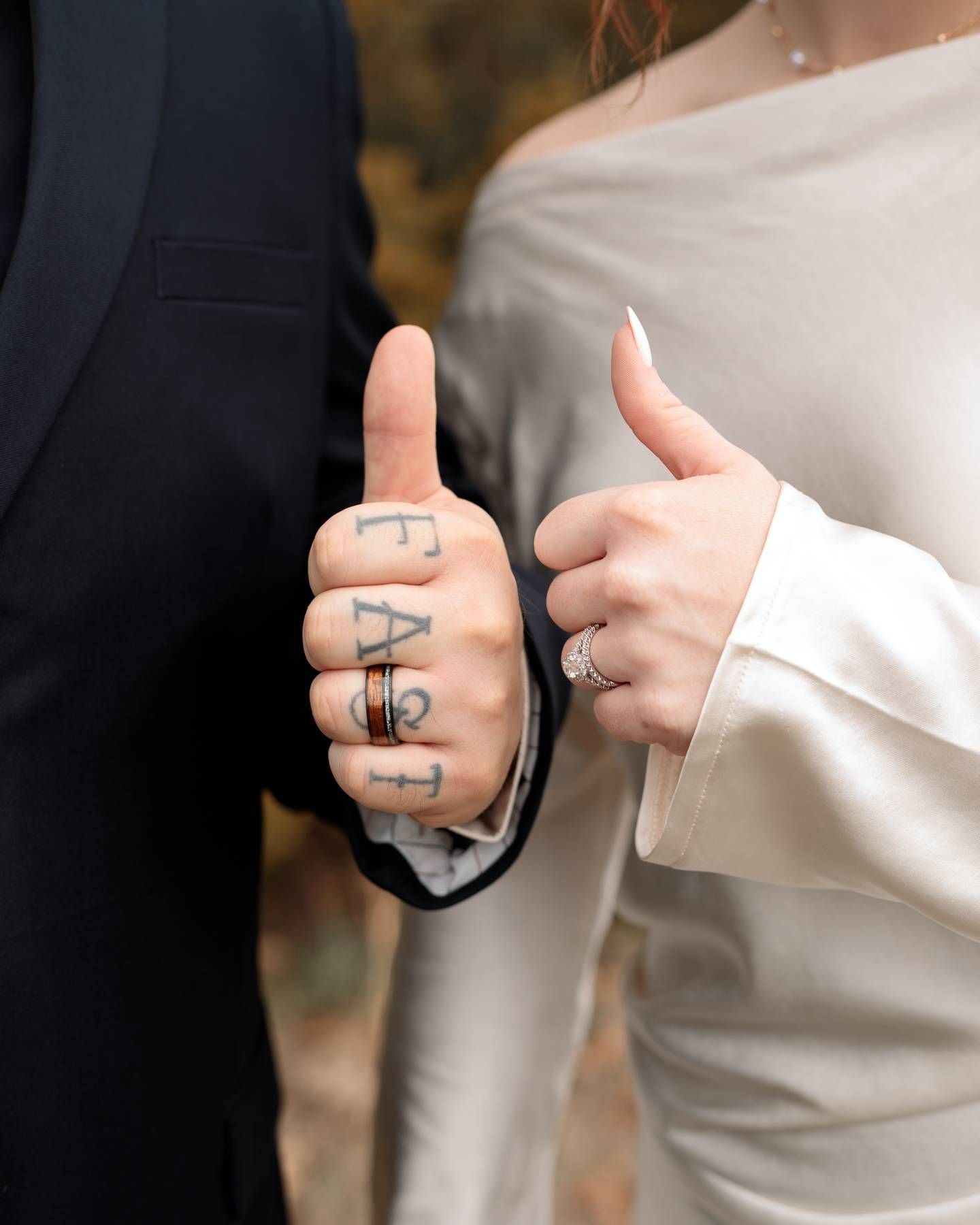 Couple gives thumbs up, groom with tattooed fingers and a ring, bride with a diamond ring, in formal wedding attire.