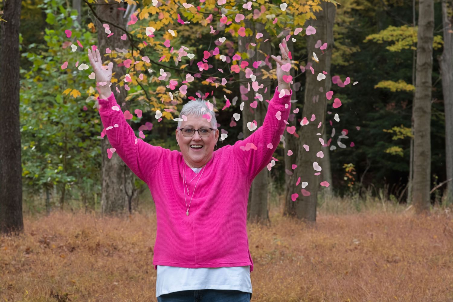 Woman in pink hoodie joyfully tossing pink and white flower petals in an autumn forest setting.