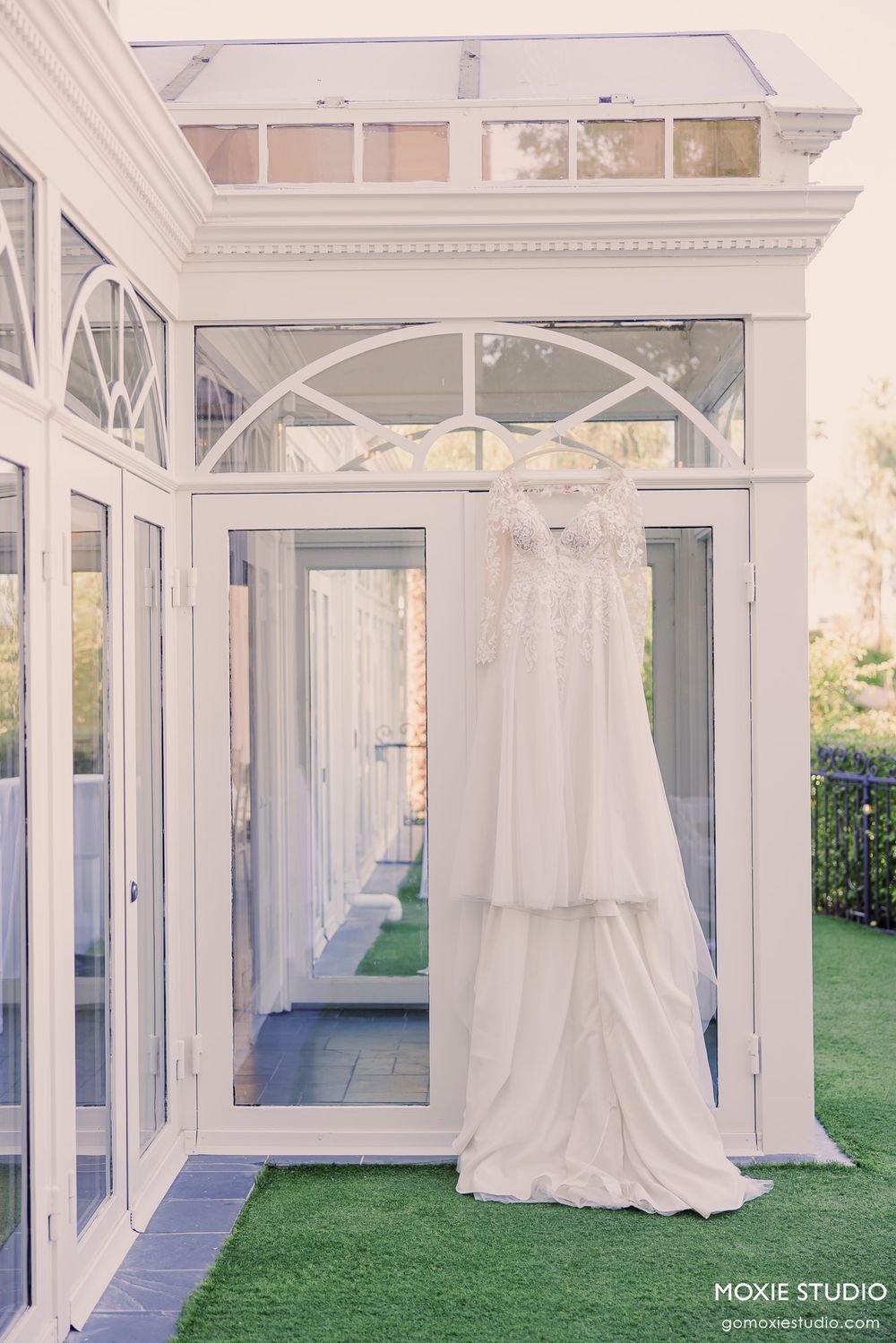 Elegant white wedding dress hanging in a bright white glass conservatory doorway with lush green turf flooring.