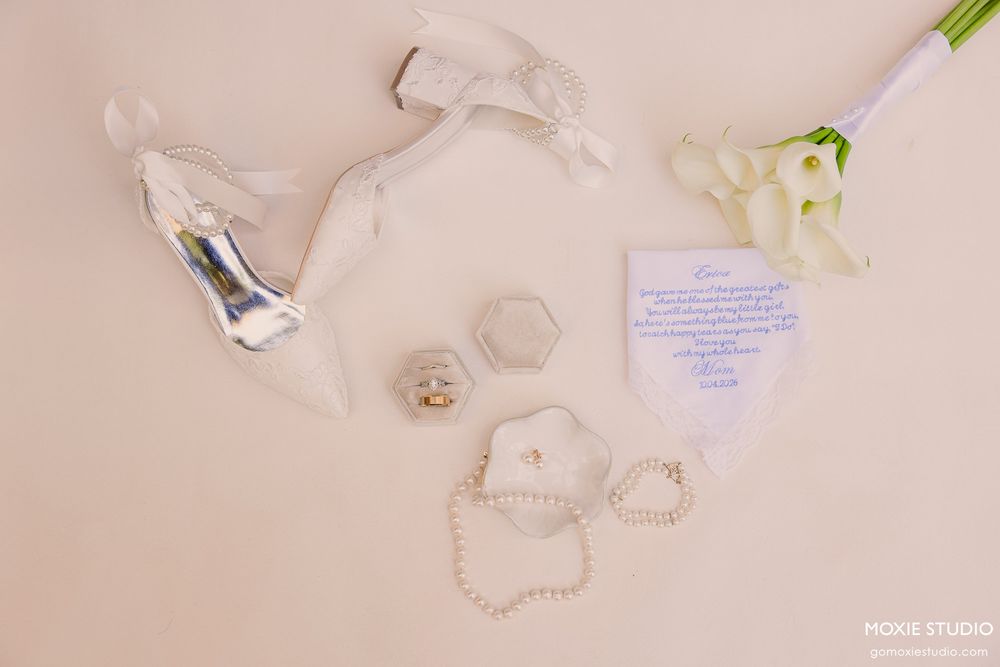 Elegant wedding flat lay featuring white heels, pearl bracelet, ring boxes, floral bouquet, and handwritten note on blush background.