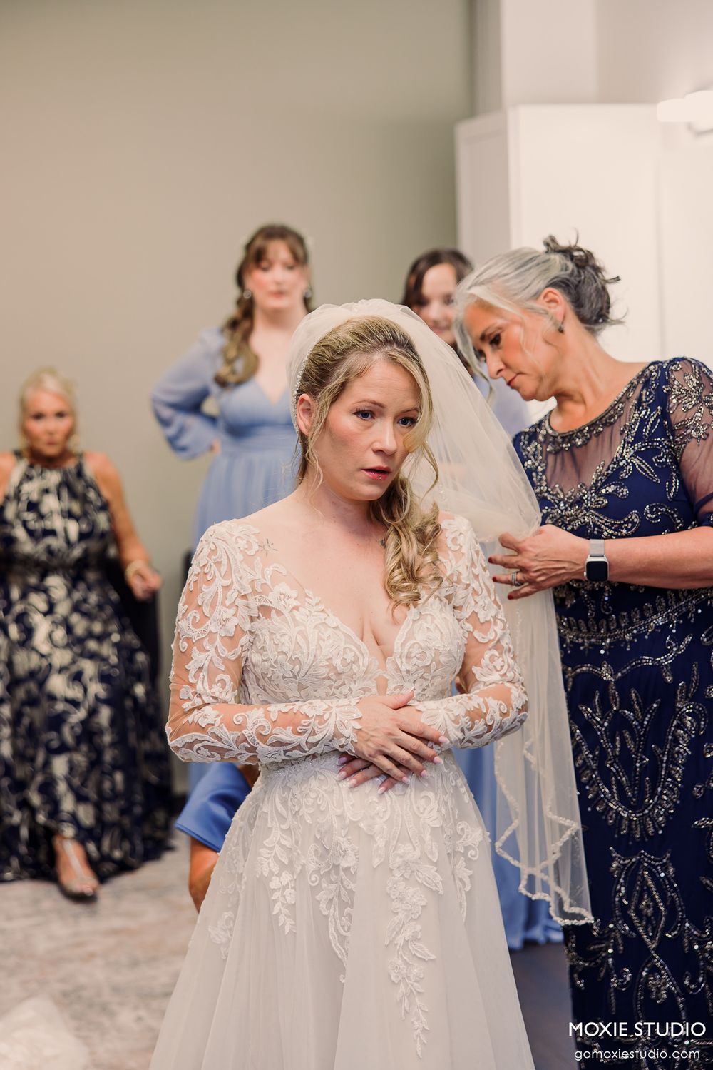 Bride in lace wedding gown and veil being helped by older woman in navy dress, bridesmaids in blue gowns in background.