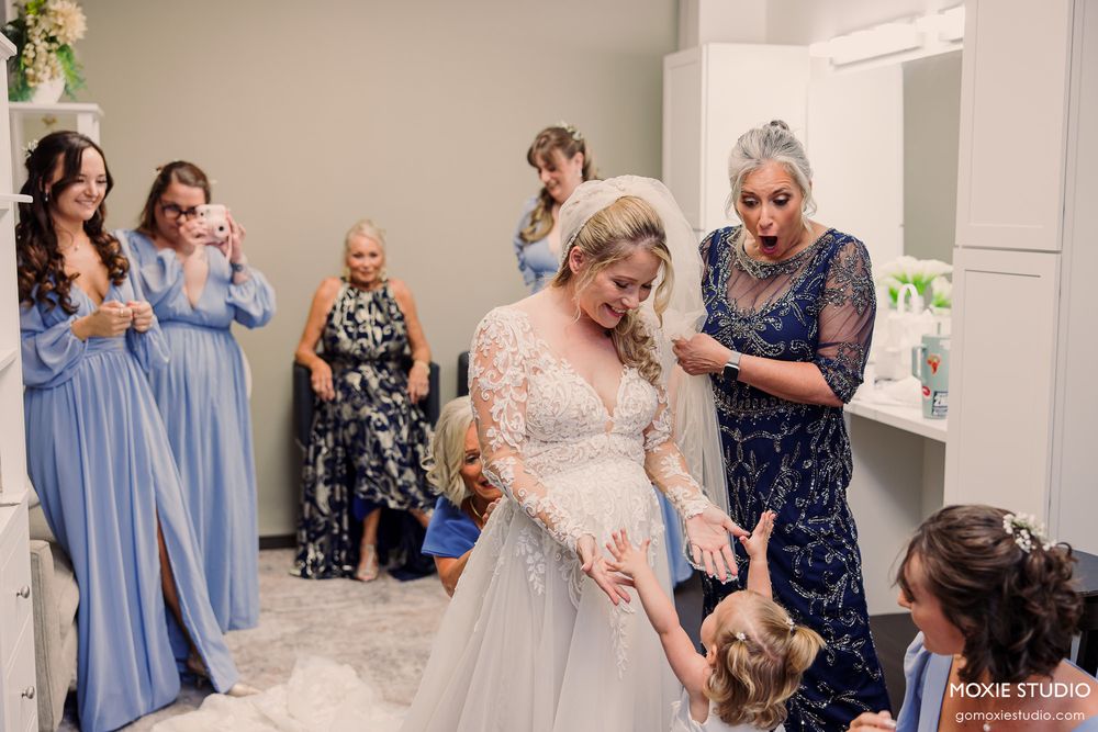 Bride in lace wedding gown surrounded by bridesmaids in blue dresses and family during bridal preparations indoors.