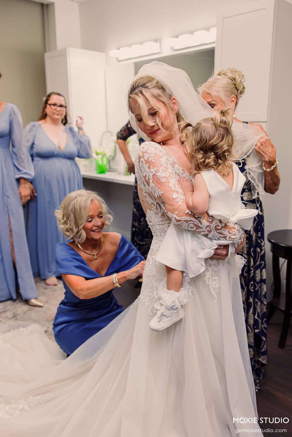 Bride in lace gown holds toddler while women in blue dresses assist with veil in bright bridal suite.