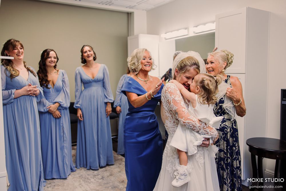 Bride embracing a child surrounded by bridesmaids in blue dresses and family members in a bright wedding preparation room.