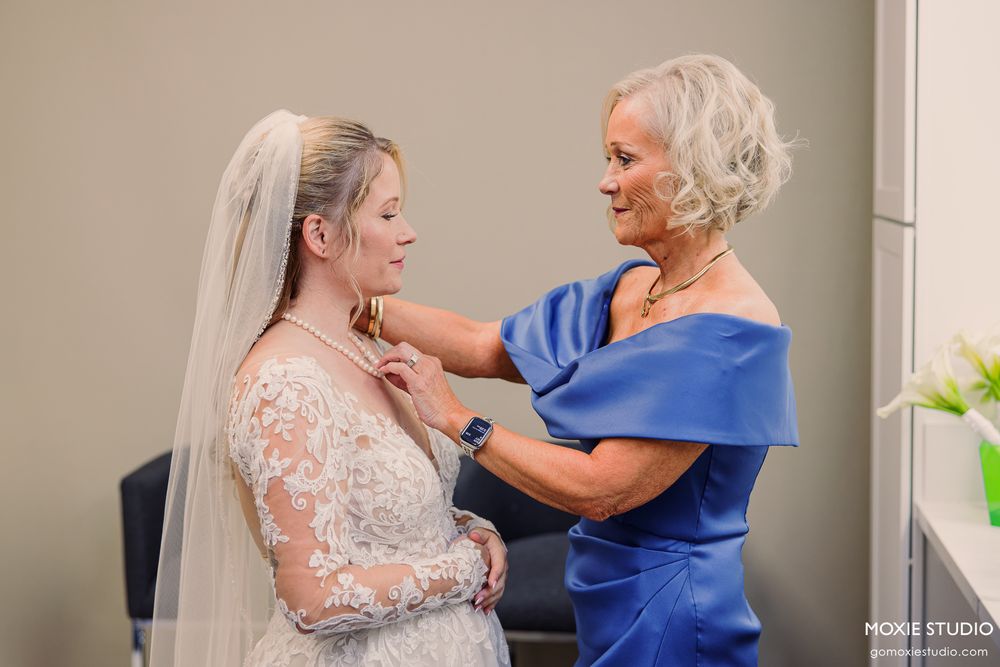 Bride in lace wedding gown and veil gets necklace fastened by older woman in blue satin dress.
