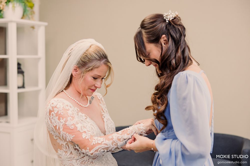 Bride in lace gown and veil shares a tender moment with bridesmaid in blue dress during wedding preparations.