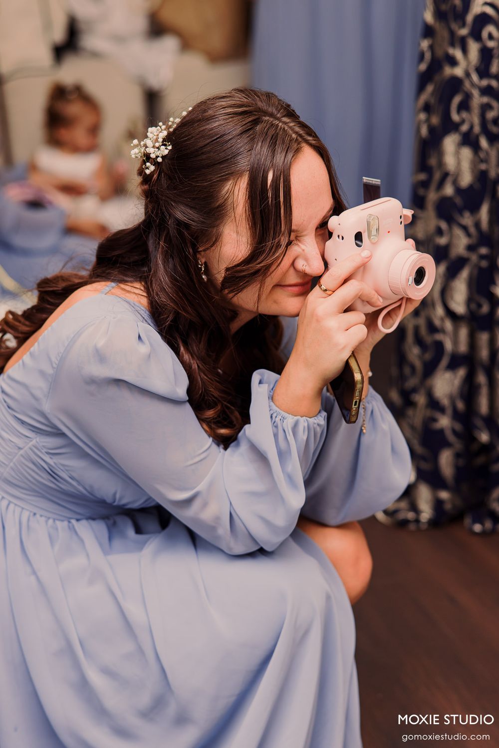 A woman in a light blue dress photographs with a pink Instax camera, wearing floral hair accessories at a wedding.