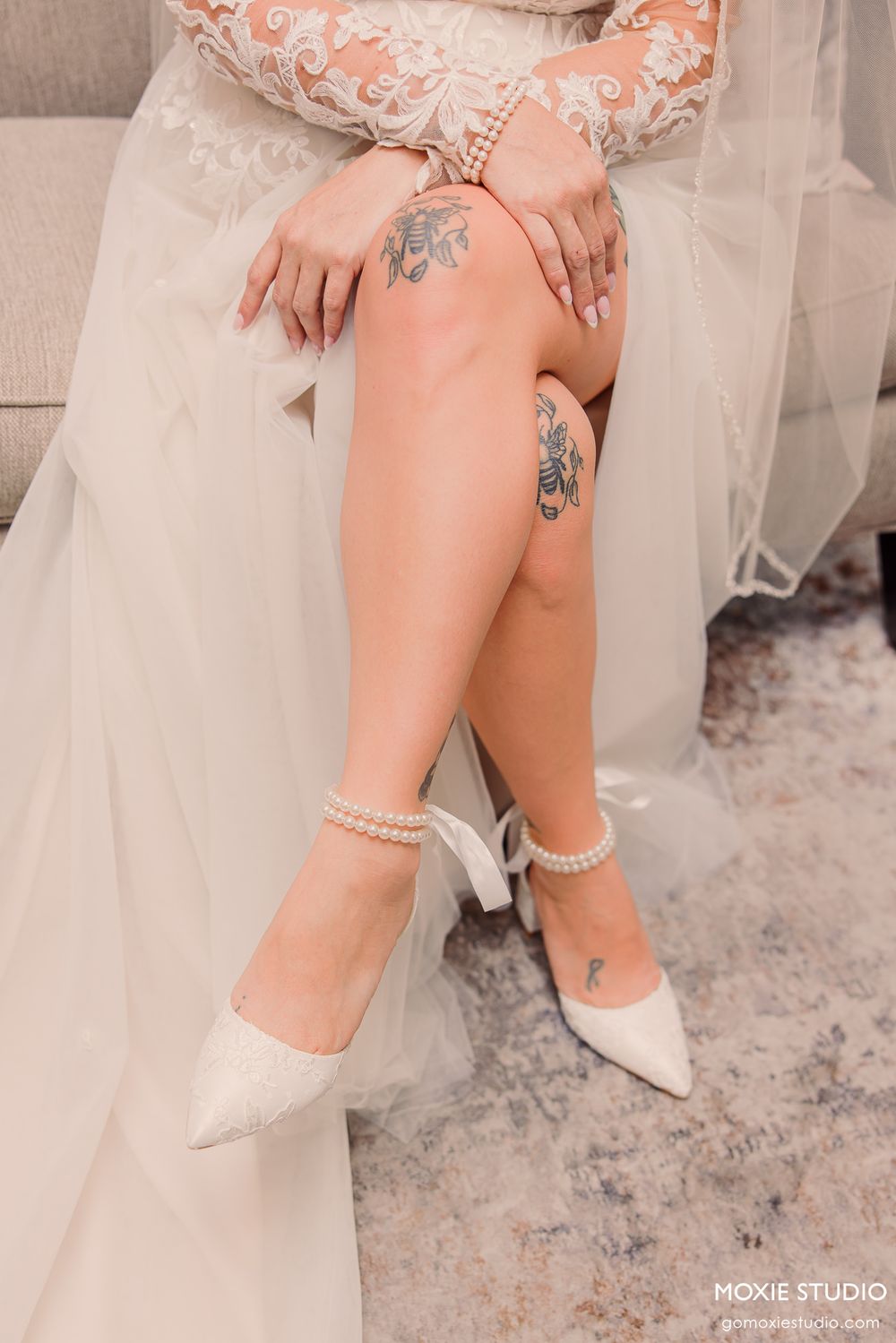 Bride in lace wedding dress showing tattooed legs and pearl ankle strap heels while seated on a sofa.