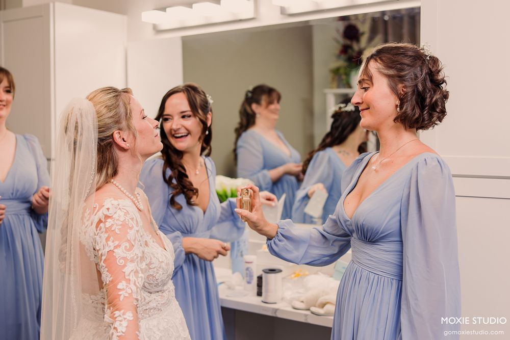 Bride laughing with bridesmaids in blue dresses during wedding preparations in a bright dressing room.