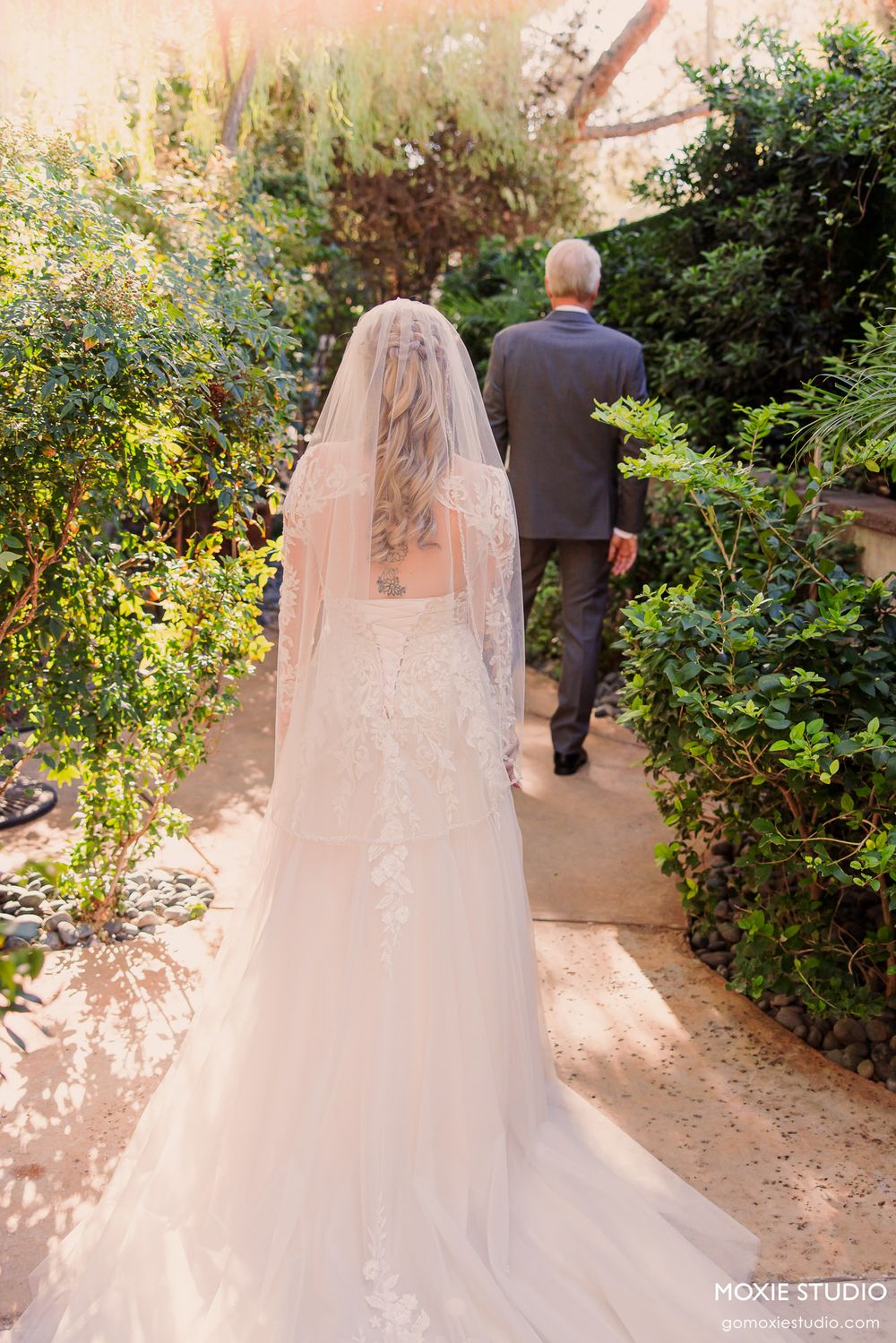 Bride in lace gown and veil walks with suited man through lush garden path on wedding day.