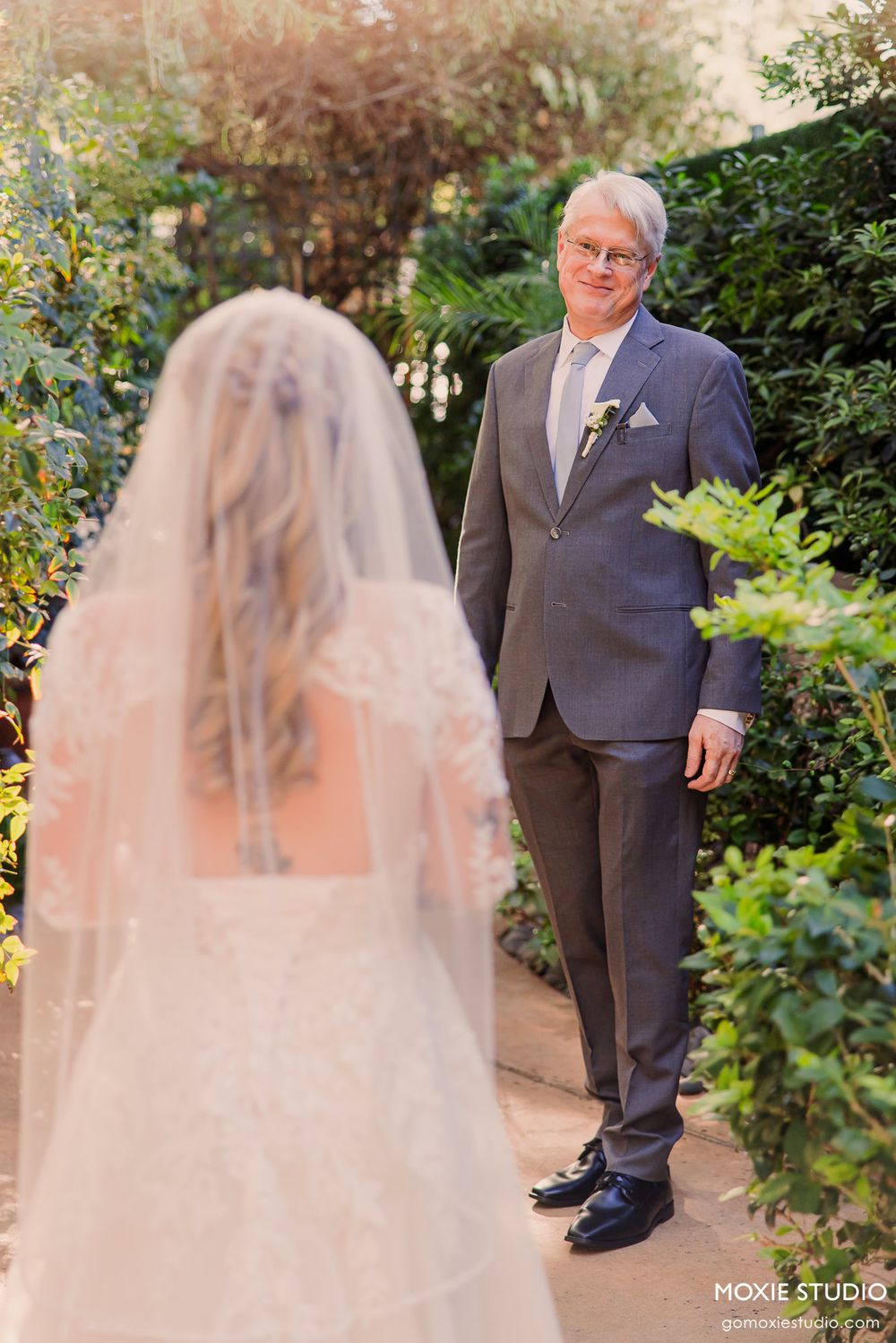 Father in gray suit smiles at bride in white lace gown and veil during outdoor first look at wedding.