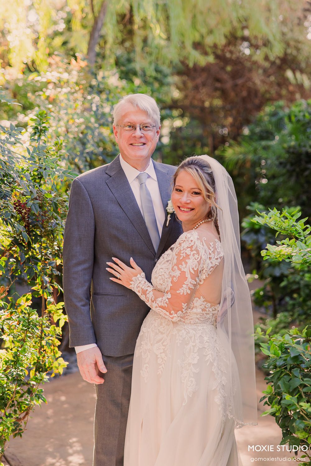 Bride in lace gown and veil poses with older gentleman in gray suit in a lush garden wedding setting.