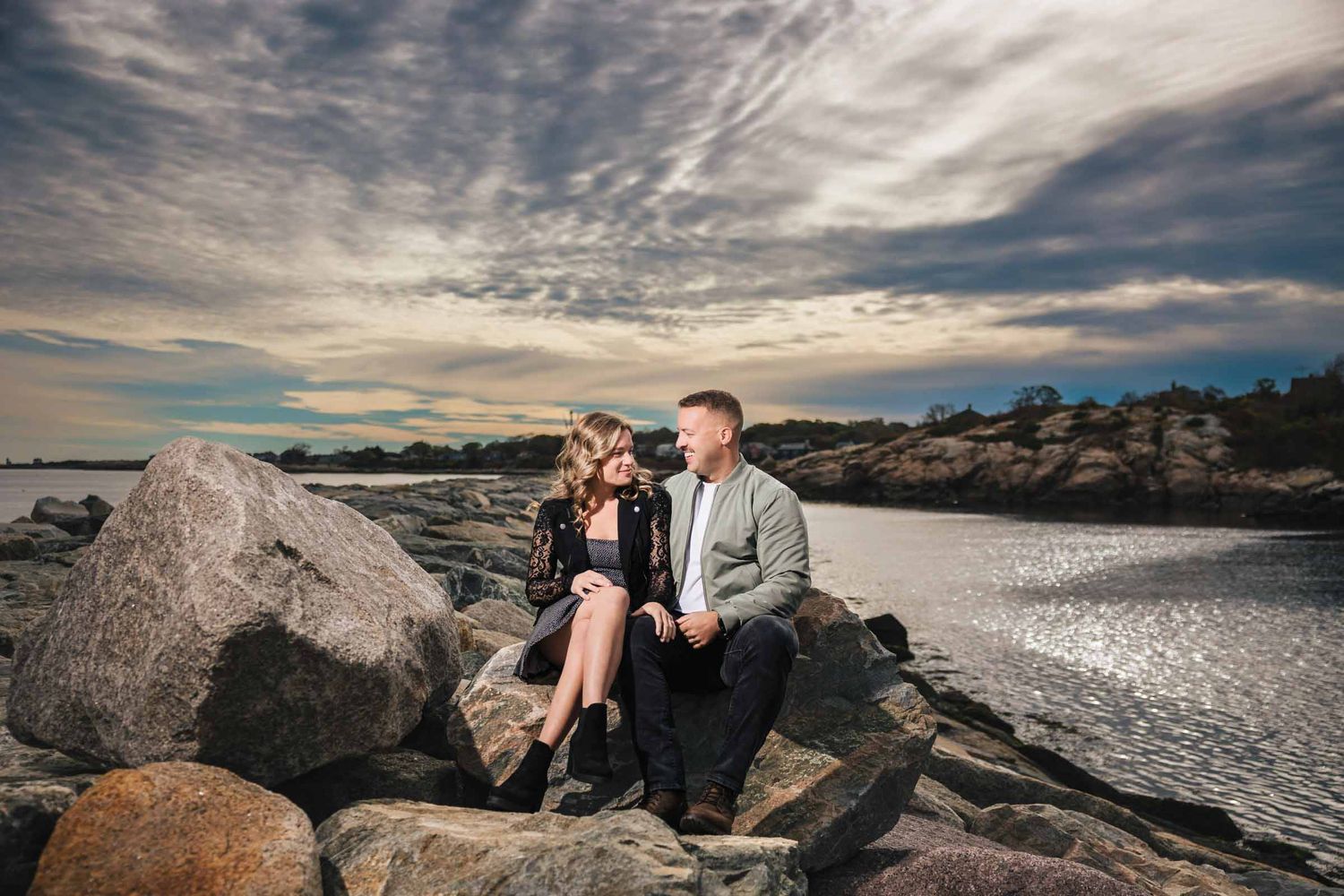Couple sitting together on rocky breaker, gazing at each other with the water and sky behind them.  Engagement at Rockport Harbor, Rockport