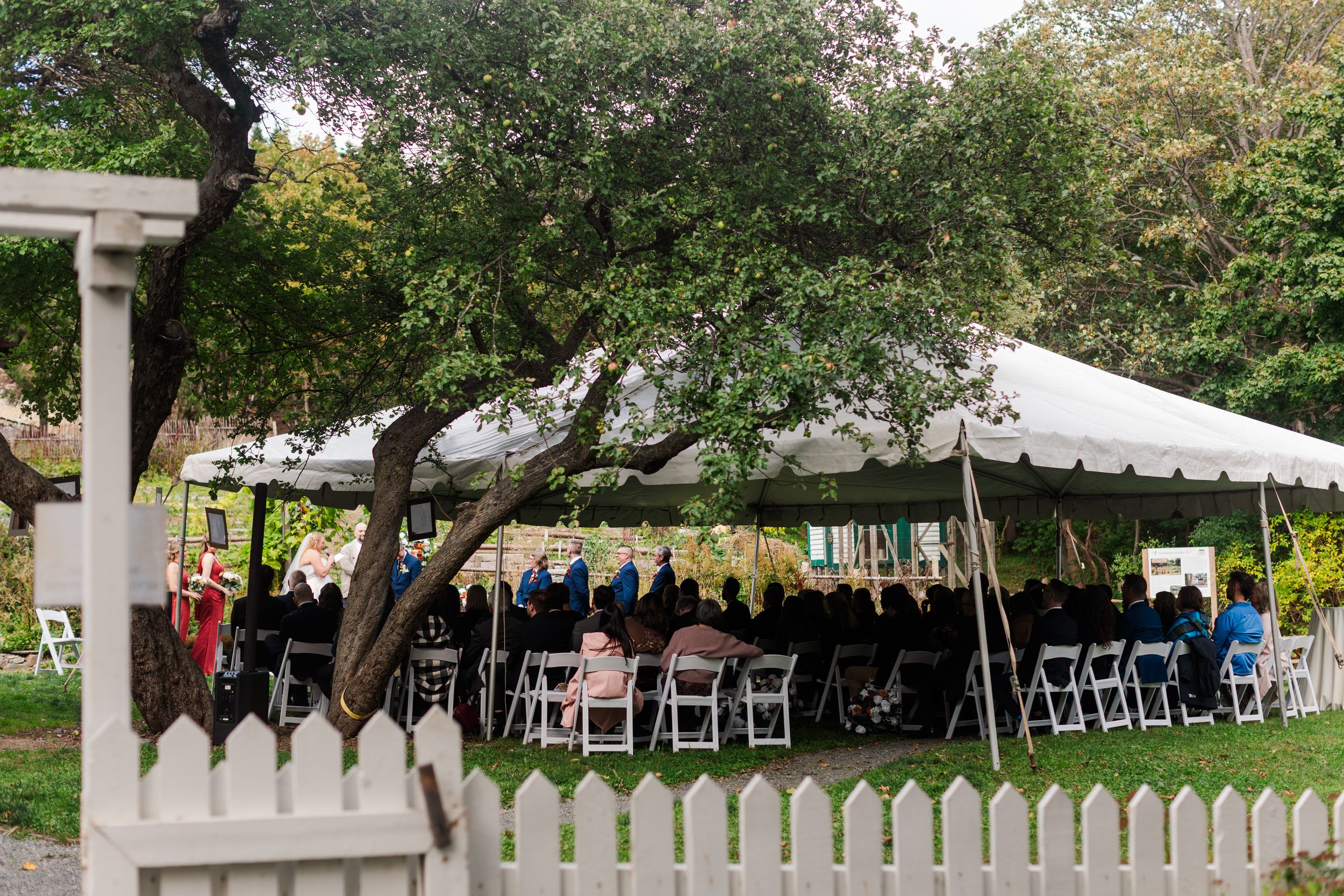 Guests seated on white chairs under a large white tent during an outdoor ceremony, framed by leafy trees and a white picket fence.