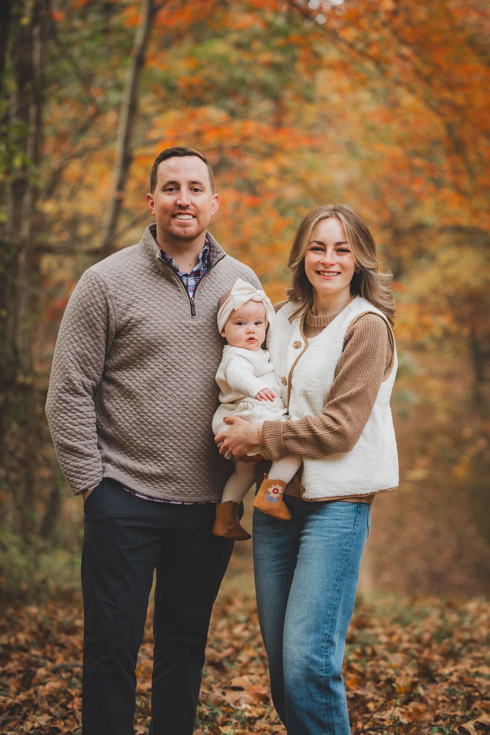 A smiling family of three poses outdoors in autumn, parents holding baby in white outfit among colorful fall foliage.