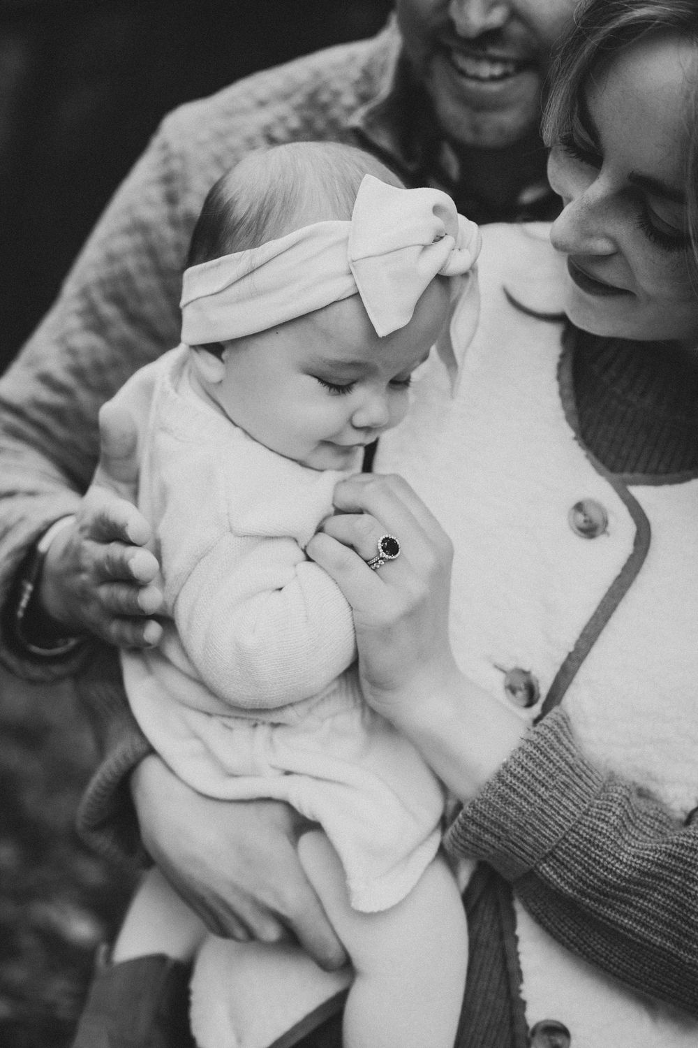 Black and white photo of smiling parents holding their adorable baby girl wearing a white bow headband.