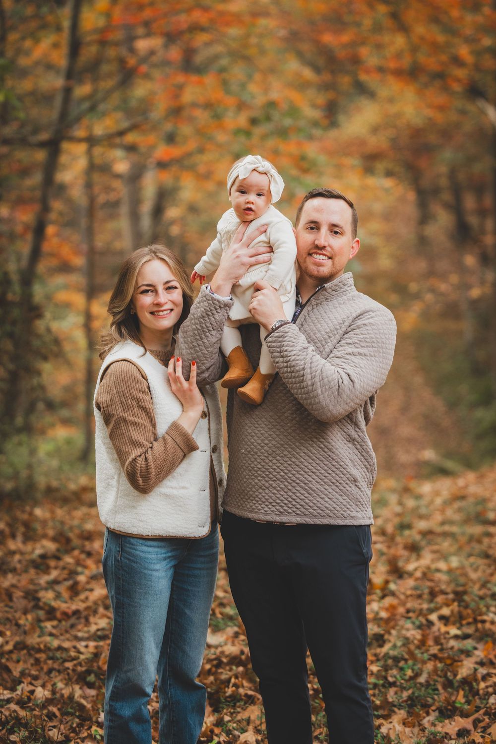 Family of three posing in autumn forest, parents holding baby in white outfit among colorful fall foliage.