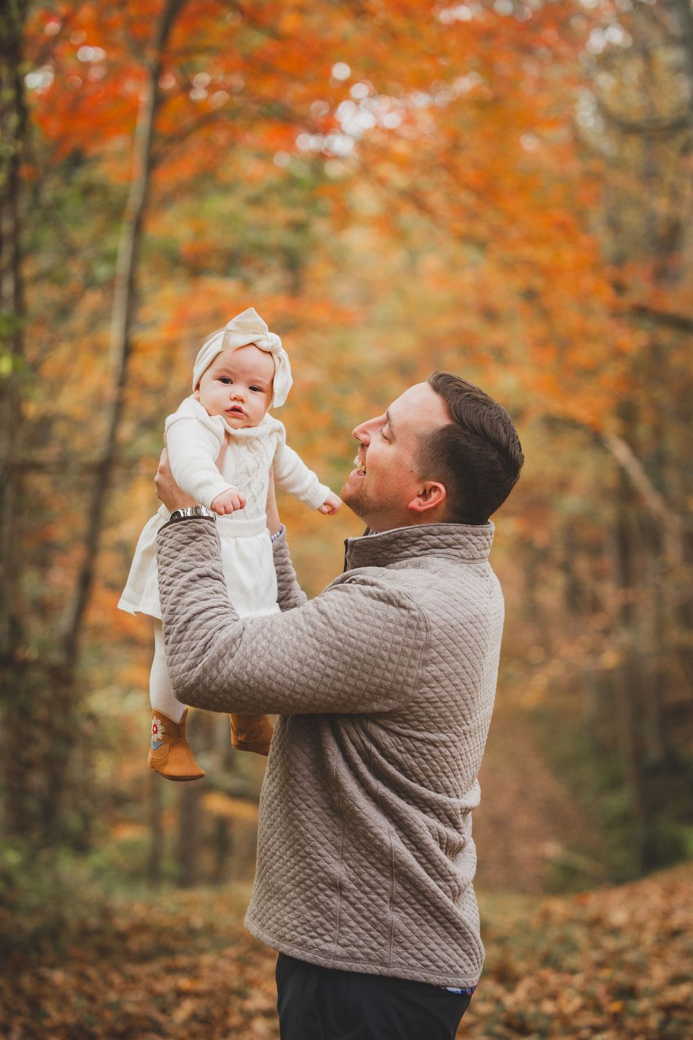Father lifting baby girl in white dress amid colorful autumn forest trees, sharing a joyful moment outdoors.