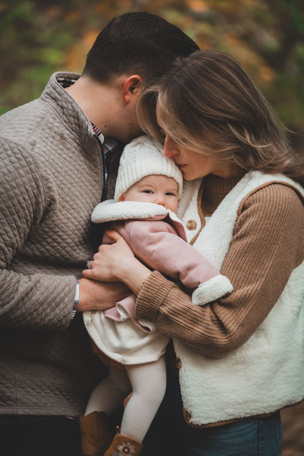 A couple tenderly kisses their newborn baby outdoors in autumn, dressed in cozy neutral sweaters.