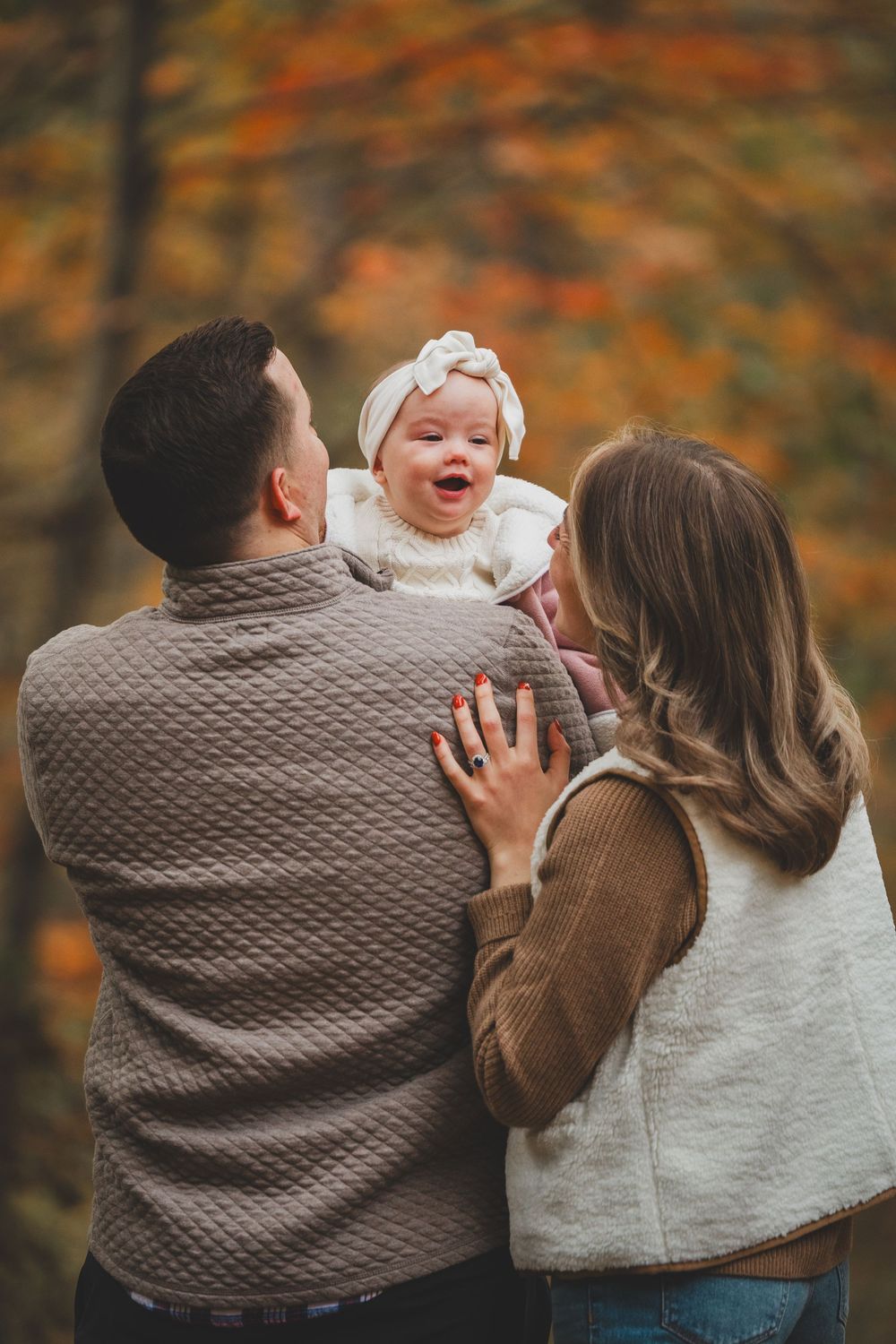 A happy baby laughs while being held by parents in a fall forest setting with colorful autumn foliage.
