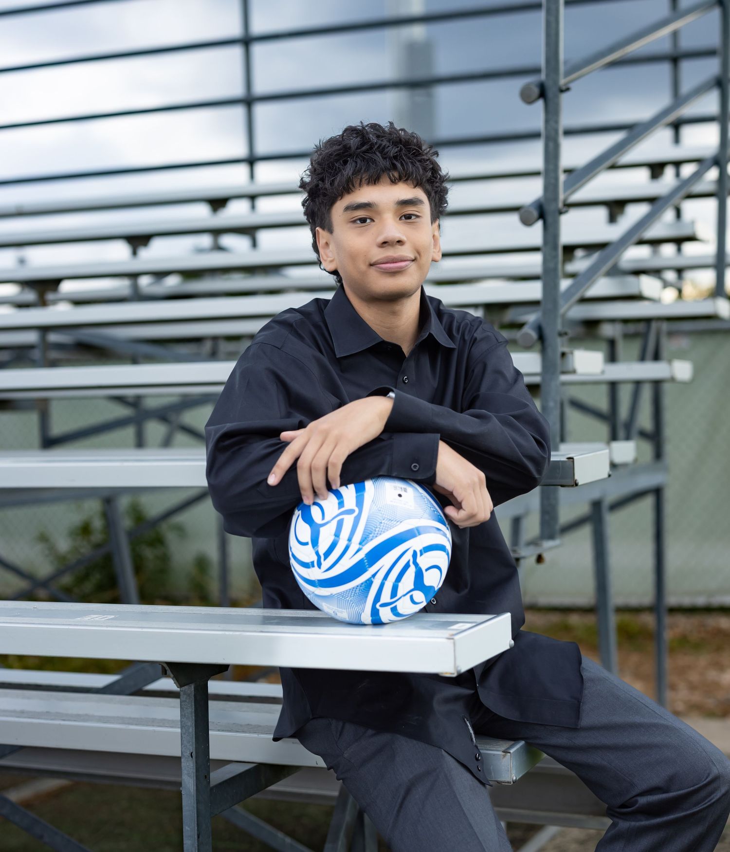 Young man in black outfit sitting on bleachers with a blue and white soccer ball.