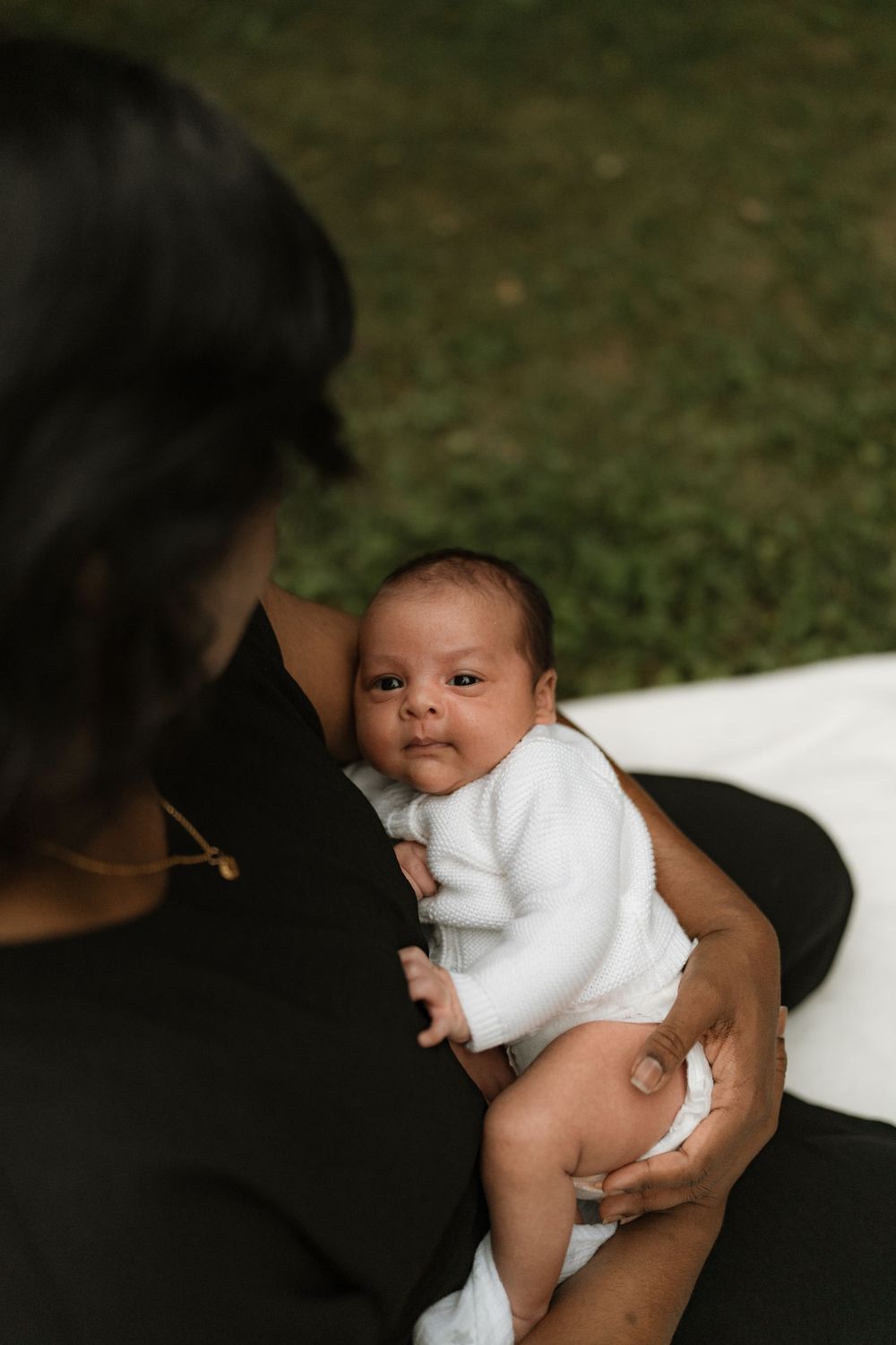 A wide-eyed newborn baby in a white outfit rests in a parent's arms on a blanket outdoors, with soft green grass in the background.