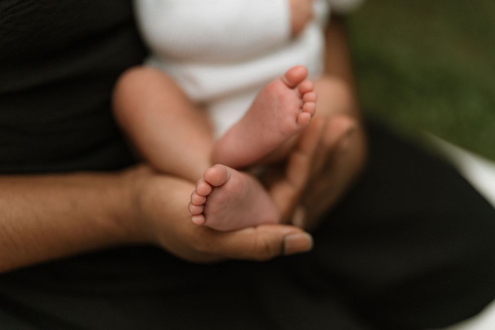 Adult hands gently cradling the tiny bare feet of a newborn baby, with soft blurred greenery in the background.