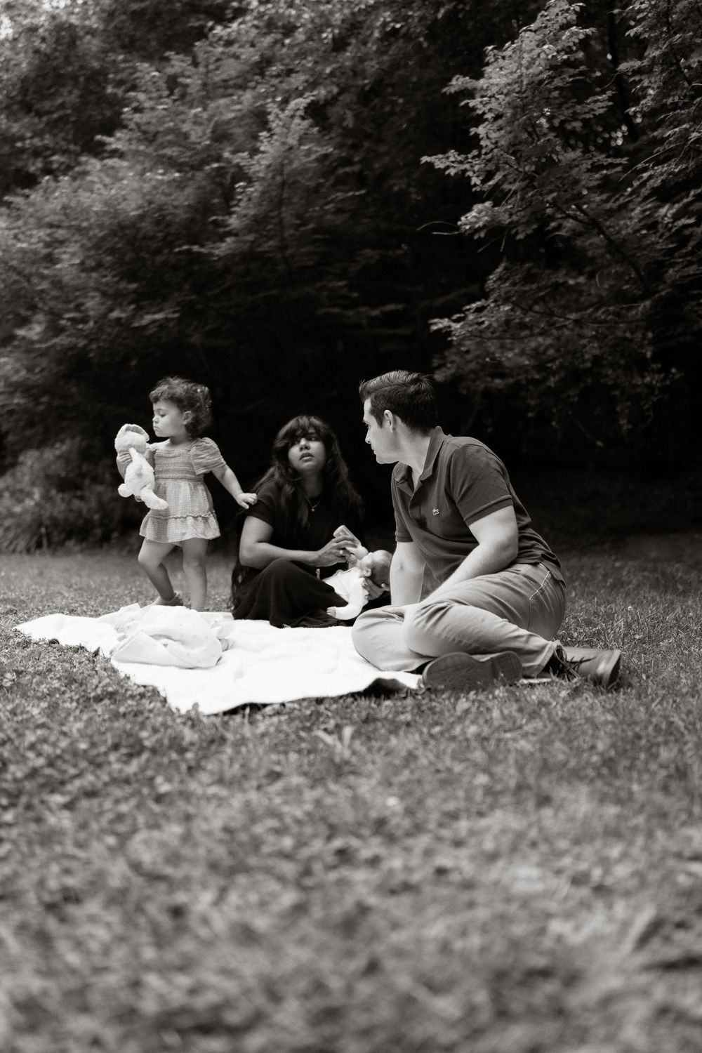Black and white photo of a family having a picnic on a blanket in a grassy park, with a toddler holding a stuffed toy near tall trees.
