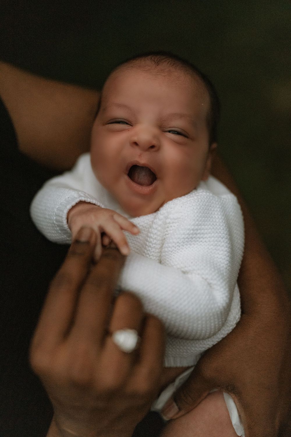 Newborn baby in a white knit sweater yawns with eyes half open while being gently held in an adult's hands, one wearing a silver ring.