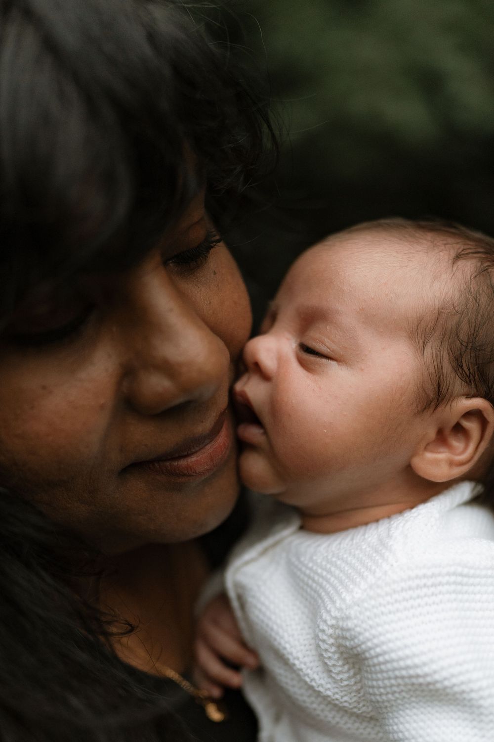 A smiling woman with long dark hair gently kisses the cheek of a sleeping newborn baby dressed in a white knit sweater.