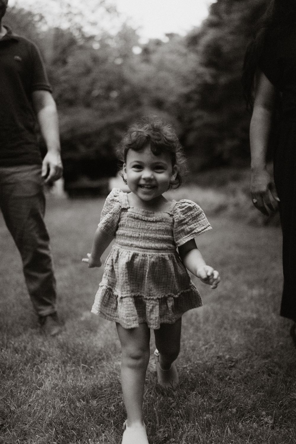 Black and white photo of a smiling toddler with curly hair running across a grassy park while two adults stand nearby.