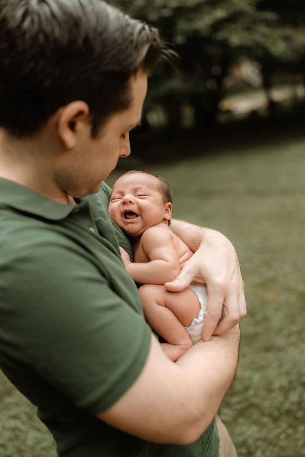 A father in a green polo shirt gently cradles a sleeping newborn baby in his arms outdoors on a grassy lawn surrounded by trees.